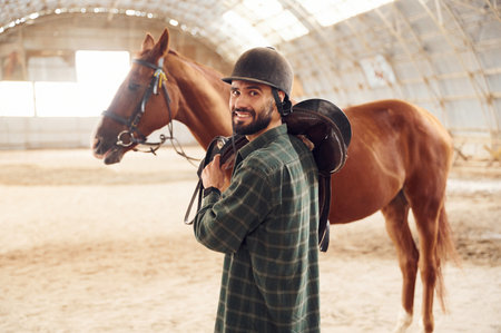 Holding the saddle in hands. Young man with a horse is in the hangar.の写真素材