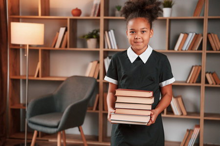 Holding books in hands. Cute african american girl in school uniform is at home library.