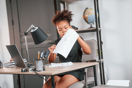 Holding paper. Cute female african american student child is sitting by the laptop.