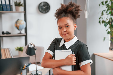Holding notepad and smiling. Cute little girl in school uniform is standing indoors in domestic room.の写真素材
