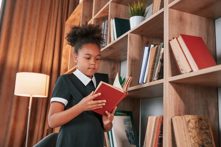 Standing and reading the book. Cute african american girl in school uniform is at home library.の写真素材