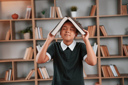 Having fun, book is on the head. Cute african american girl in school uniform is at home library.