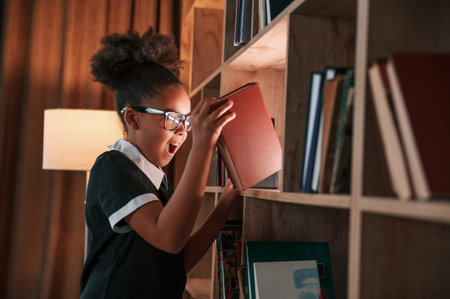 With book in hand. Beautiful light. Cute african american girl in school uniform is at home library.