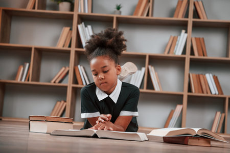 Sitting by the table with books. Cute african american girl in school uniform is at home library.