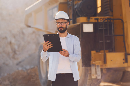 In glasses, reading documents. Man in uniform is working in the quarry at daytime.