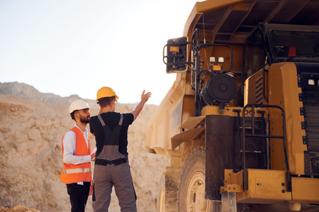 Looking at big haul truck. Two men in uniform are working together in the quarry.
