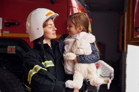 Kid is holding bunny toy. Firefighter woman in uniform is with a little girl.