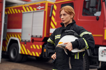 Heavy truck behind. Holding protective helmet. Woman firefighter in uniform is at work in...
