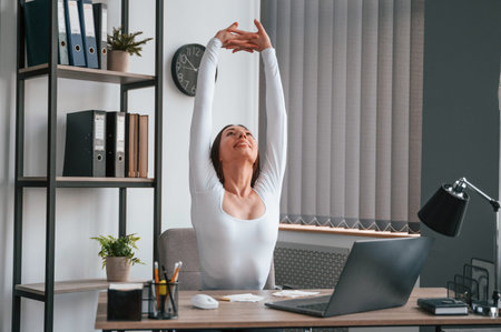 Takes a break, stretches her hands. Young beautiful woman in white clothes is in the office.