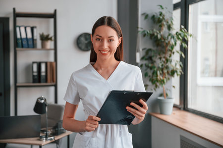 Smiling young beautiful woman in white clothes is in the office with notepad.の写真素材