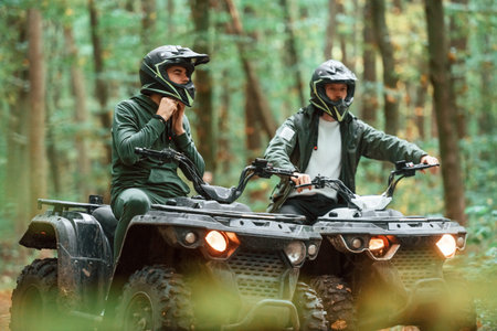 Wearing helmets. Two male atv riders is in the forest together.