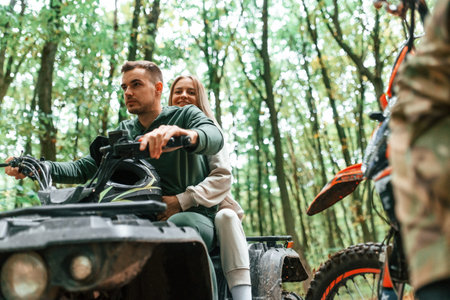 Lovely couple is sitting on the ATV with man on motorcycle in the forest.