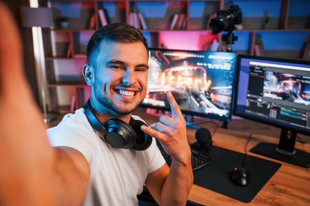 Making selfie, celebrating victory. Male game streamer in casual clothes is indoors with pc.