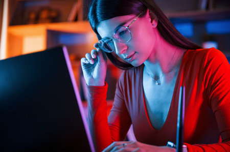 Focused on the work. Beautiful woman in glasses and red wear is sitting by the laptop in dark room...