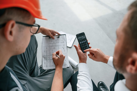 Sitting, using calculator. Manager with foreman are working on the construction site together.