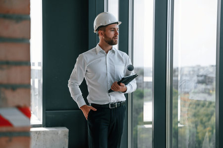 Standing near panoramic windows. Man in white shirt is working indoors at construction site....