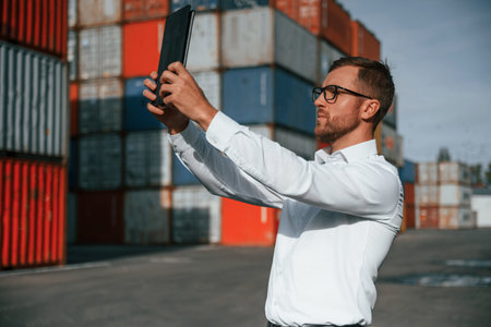 With modern tablet. Making a photos of objects. Male worker is on the location with containers.