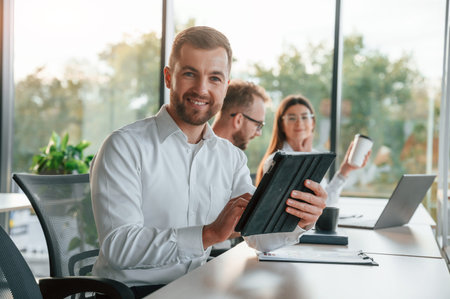 Man is looking at the camera. Three people in formal clothes are working in the modern office together.の写真素材