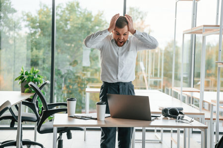 Angry boss is standing by the table with laptop on it. Man in formal clothes is working in the...