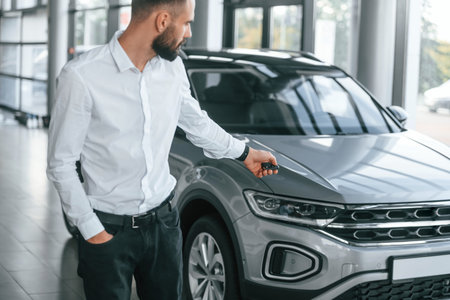 Using the keys remote controller. Young man in white clothes is in the car dealership.
