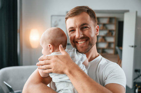Standing with baby in hands. Father with toddler is at home, taking care of his son.