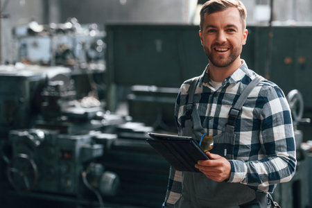 Standing and using graphic tablet. Factory male worker in uniform is indoors.