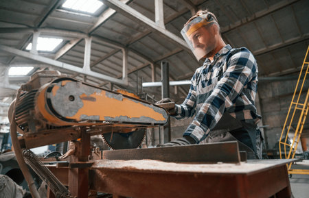 Cutting the wood by saw. Factory male worker in uniform is indoors.