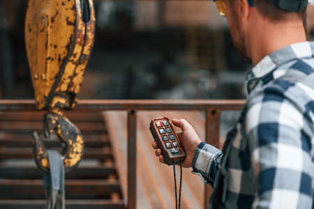 Using the controller with buttons to operate the little crane machine. Factory male worker in uniform is indoors.の写真素材