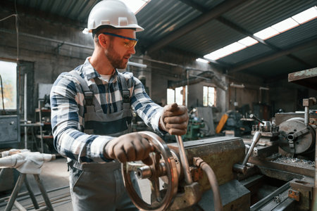 Factory male worker in uniform is indoors. In defensive glasses and hard hat.