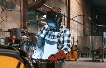 In protective mask. Welding the iron. Factory male worker in uniform is indoors.