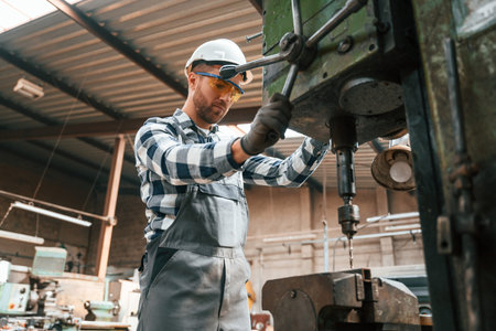 Factory male worker in uniform is indoors. Operating the drill machine.