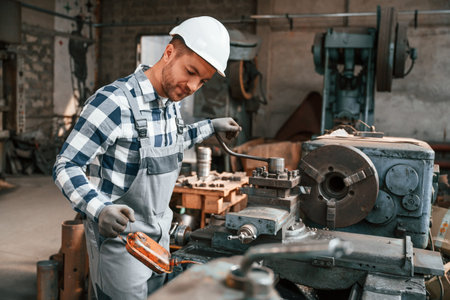 Handyman is working with factory machine. Warehouse employee in uniform is indoors.