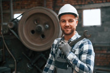 Smiling and holding wrench. Factory male worker in uniform is indoors.