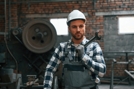 Standing and showing wrench. Factory male worker in uniform is indoors.