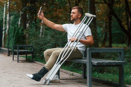 Using smartphone to make selfie. Man with crutches is in the park outdoors. Having leg injury.