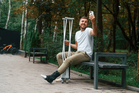 Using smartphone to make selfie. Man with crutches is in the park outdoors. Having leg injury.