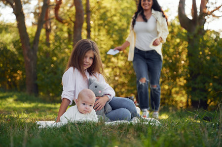 Kid is sitting with her little brother on the ground with mother behind. In the forest. Having weekend.の写真素材