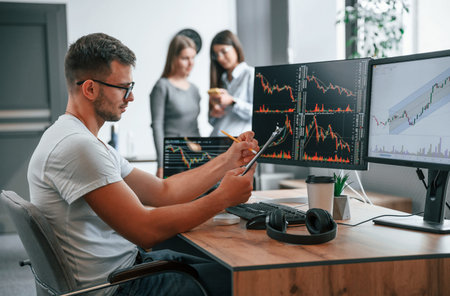 Stock broker is sitting by computer and working with exchange technology. Women is standing and...