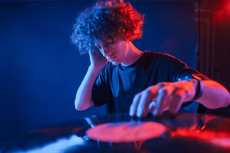 Dj is playing music from vinyl. Young man with curly hair is indoors illuminated by neon lighting.の写真素材