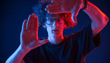 Gesture of making photo, pretending. Young man with curly hair is indoors illuminated by neon lighting.の写真素材