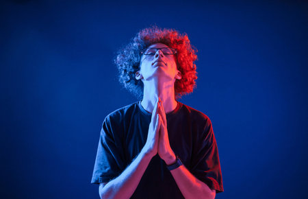 Holding hands in praying position. Young man with curly hair is indoors illuminated by neon lighting.の写真素材