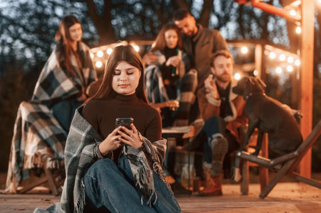 Woman with smartphone is sitting. Group of people is spending time together on the backyard at evening time.の写真素材