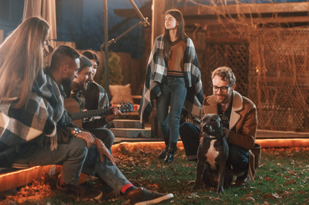 Man is petting his dog. Group of people is spending time together on the backyard at evening time.の写真素材