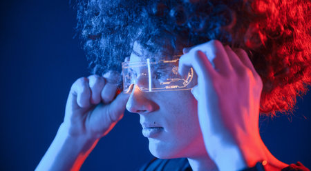 Wearing the futuristic glasses. Young man with curly hair is indoors illuminated by neon lighting.の写真素材