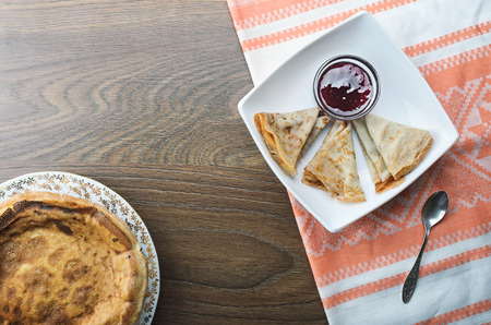 Pancakes with jam on a plate on a  wooden table covered with a rustic tablecloth. Pancakes with raspberry sauce, food closeup. Wooden table background with copy space. Top view. Russian pancakes. Pancake Tuesday.の写真素材