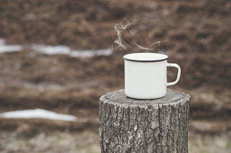 Old mug with a hot drink on a wooden stump in the outdoors. Photo in vintage color image style. Coffee break outdoors. Morning Still life. Morning at the campsite.の写真素材