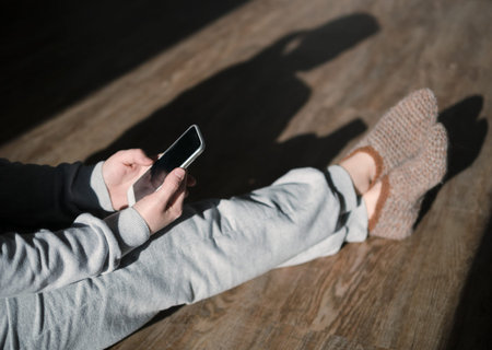 Woman sitting at cozy home interior and using modern smartphone device at sunset. Concept online...
