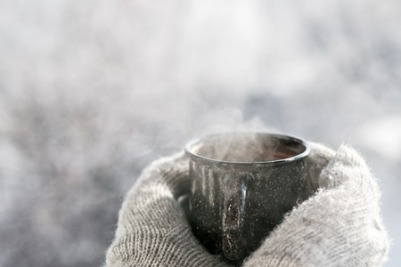 Woman hands in mittens holding a cozy steaming mug or cup with hot cocoa, tea or coffee. Beautiful girl enjoying winter morning or evening outdoors under snowfall. Winter and Christmas time conceptの写真素材