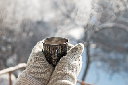 Woman hands in mittens holding a cozy steaming mug or cup with hot cocoa, tea or coffee. Beautiful girl enjoying winter morning or evening outdoors under snowfall. Winter and Christmas time conceptの写真素材