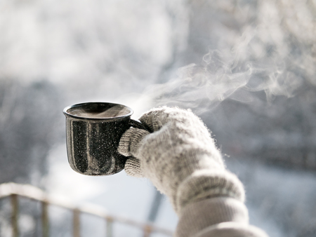 Woman hands in mittens holding a cozy steaming mug or cup with hot cocoa, tea or coffee. Beautiful girl enjoying winter morning or evening outdoors under snowfall. Winter and Christmas time conceptの写真素材